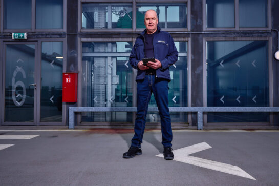 Man in TÜV Nord-jas met een tablet staat op een parkeerdek voor een modern glazen gebouw.
